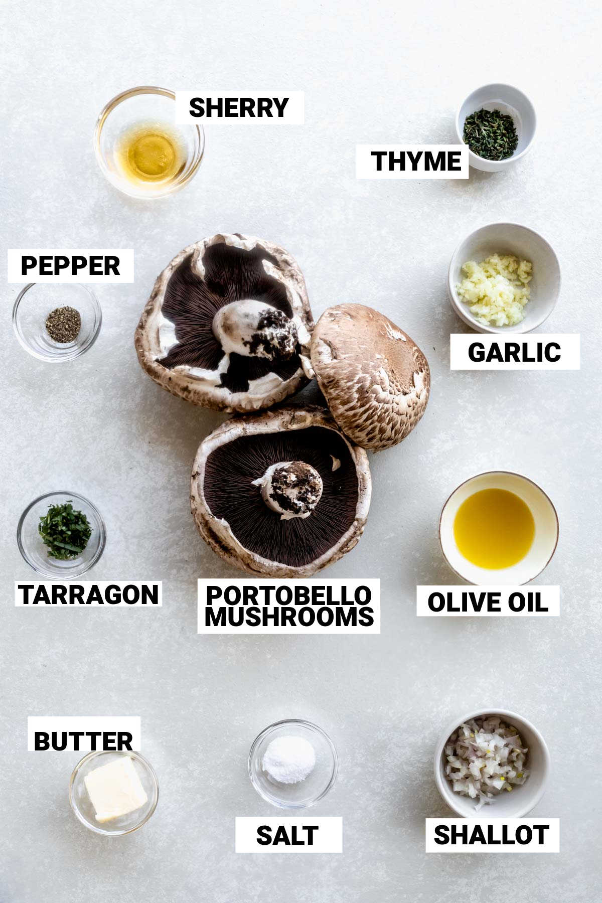 Ingredients for sautéed portobello mushrooms on a table. Mushrooms in the middle of the table and small bowls surrounding it containing garlic, oilive oil, tarragon, pepper, salt, butter, shallot, sherry, and thyme. All ingredients have a label on them saying what they are.