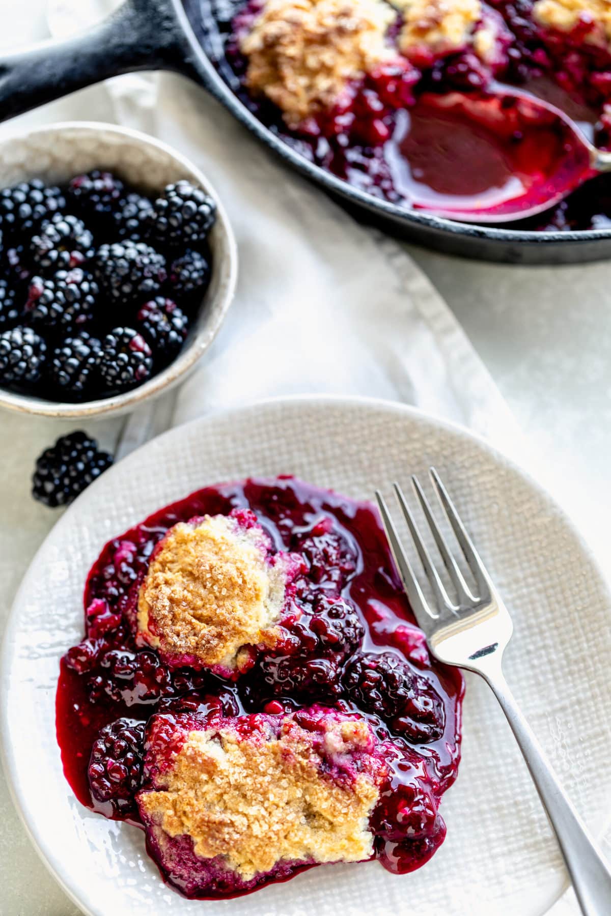 Blackberry Cobbler in a white bowl with a fork, and a small bowl of blueberries to the left hand side.