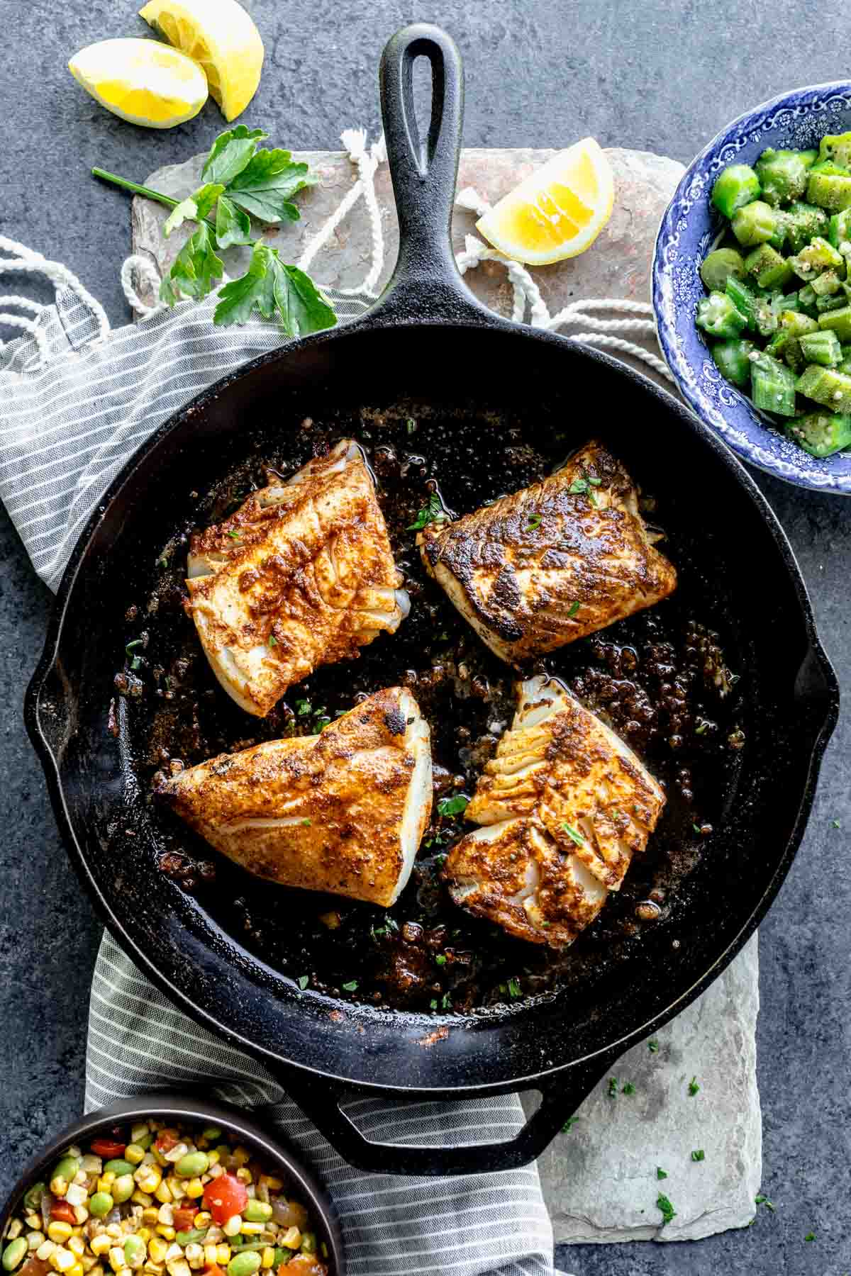 blackened cod cooking in a cast iron skillet with sides of vegetables on the counter beside it