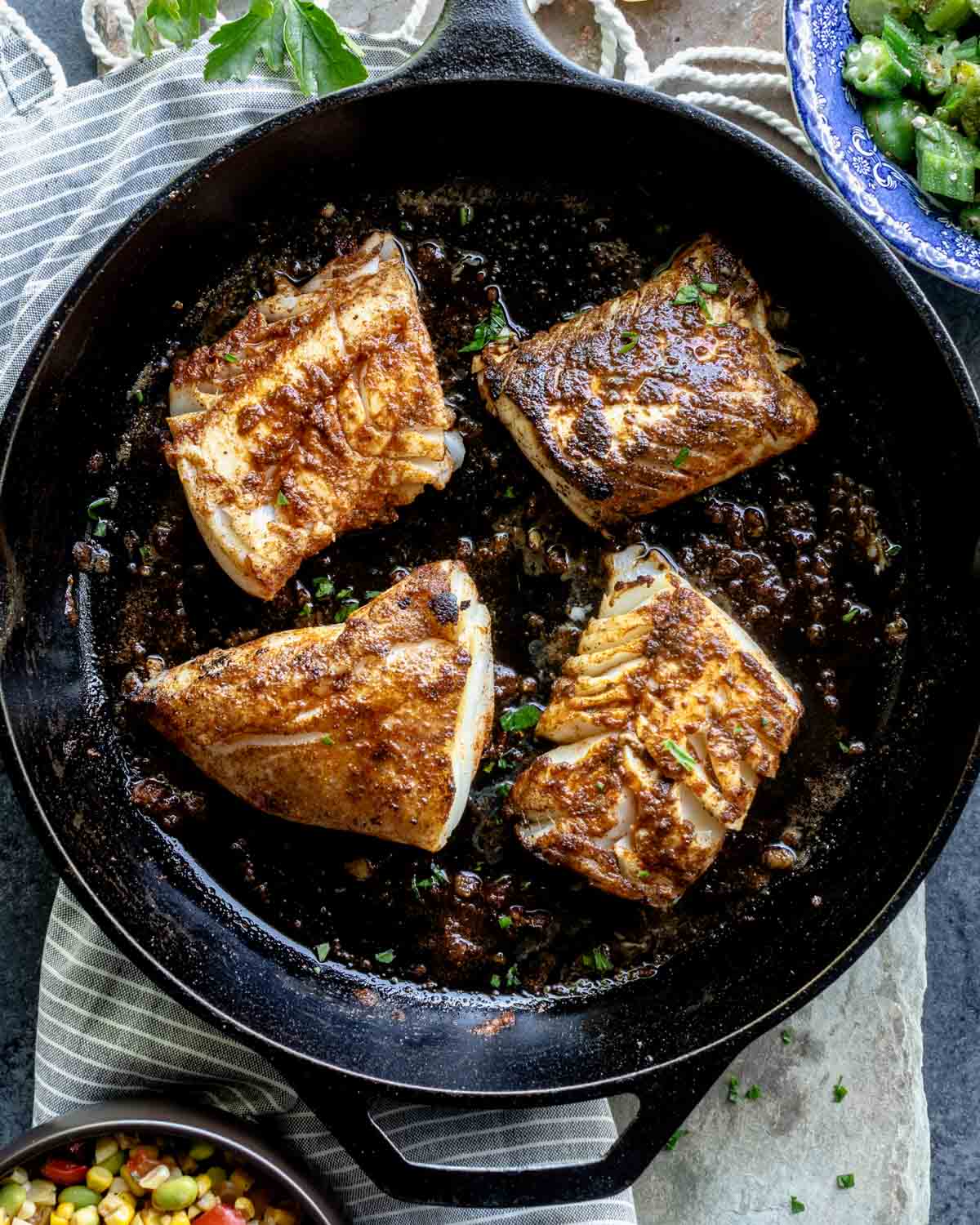 blackened cod in a cast iron skillet beside bowls of vegetables on a counter surface
