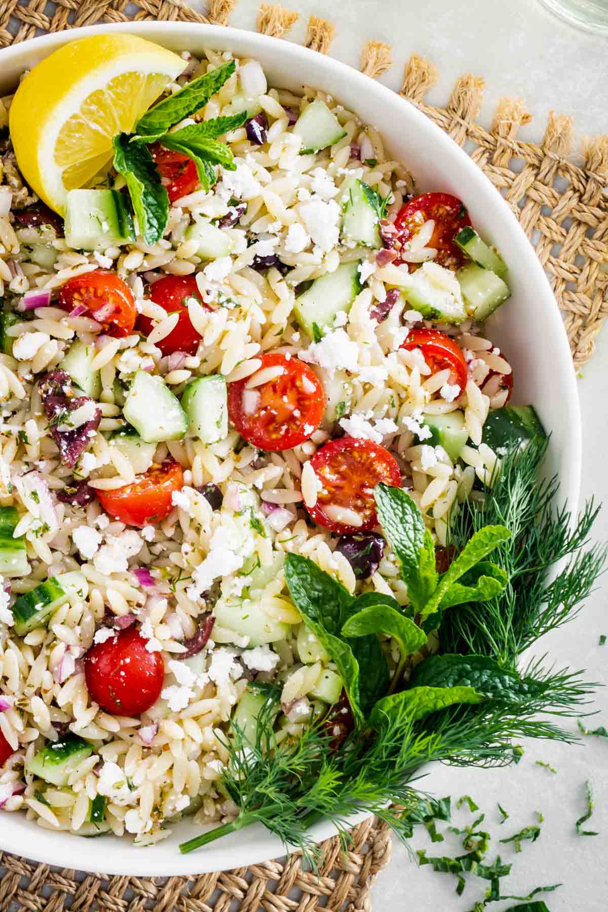 Close up of Orzo Pasta Salad in a white bowl on a table with a slice of lemon and a leafy garnish on top.