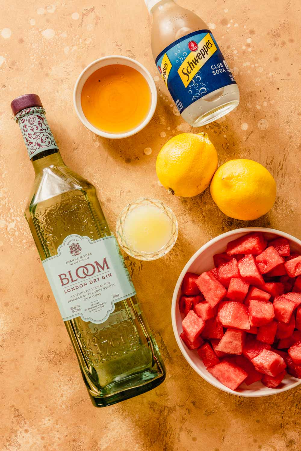 bottle of gin, two lemons, egg white, honey, club soda and diced watermelon set out on a counter