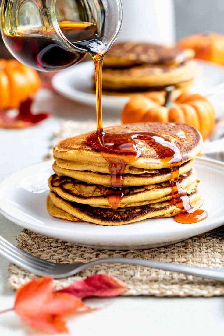 puring maple syrup over a tall stack of pumpkin pancakes with orange leaves in the foreground