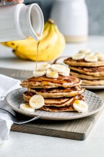 whole wheat banana pancakes stacked on a plate with banana slices and maple syrup drizzled on top