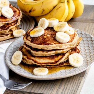 whole wheat banana pancakes stacked on a plate with banana slices and maple syrup