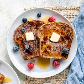 two slies of french toast on a white plate with berries, butter and syrup