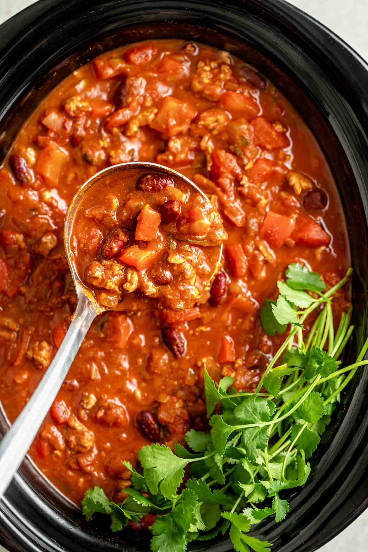 crockpot full of turkey chili with a ladle holding a close up and cilantro in the corner