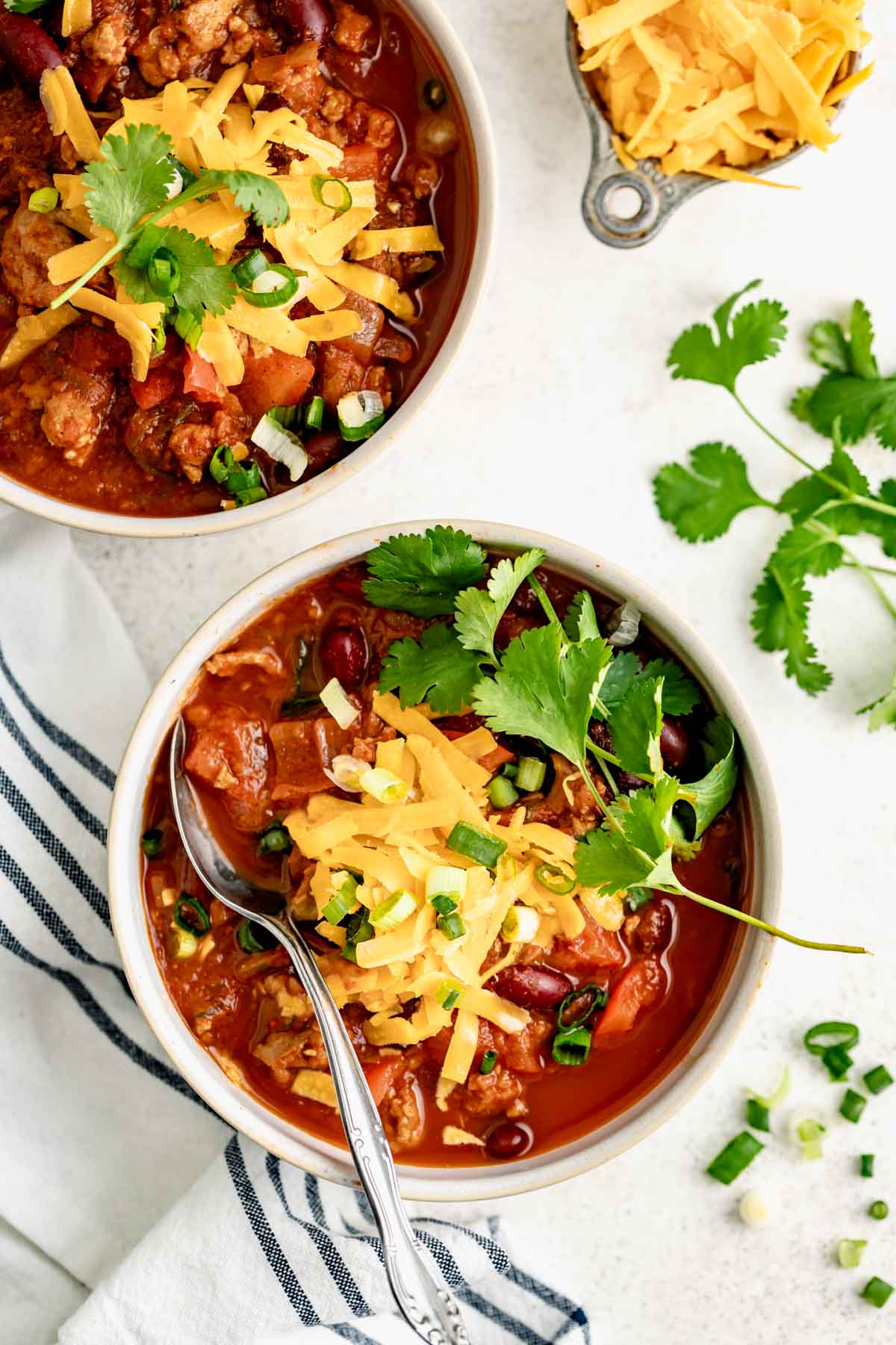 a serving bowl full of ground turkey chili covered in cheddar cheese, scallions, jalapeño, and cilantro for garnish.