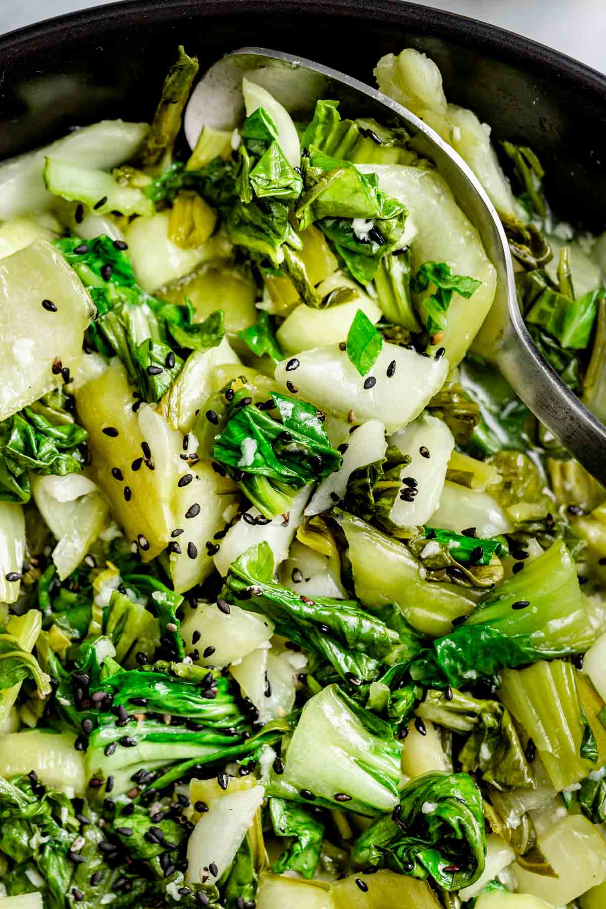 a spoon tucking into a bowl of sauteed bok choy with sesame seeds sprinkled on top