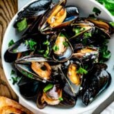 a white bowl filled with steamed mussels with parsley on top and two slices of grilled baguette on the table next to it