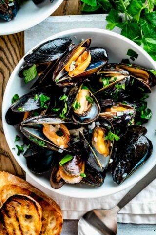 a white bowl filled with steamed mussels with parsley on top and two slices of grilled baguette on the table next to it