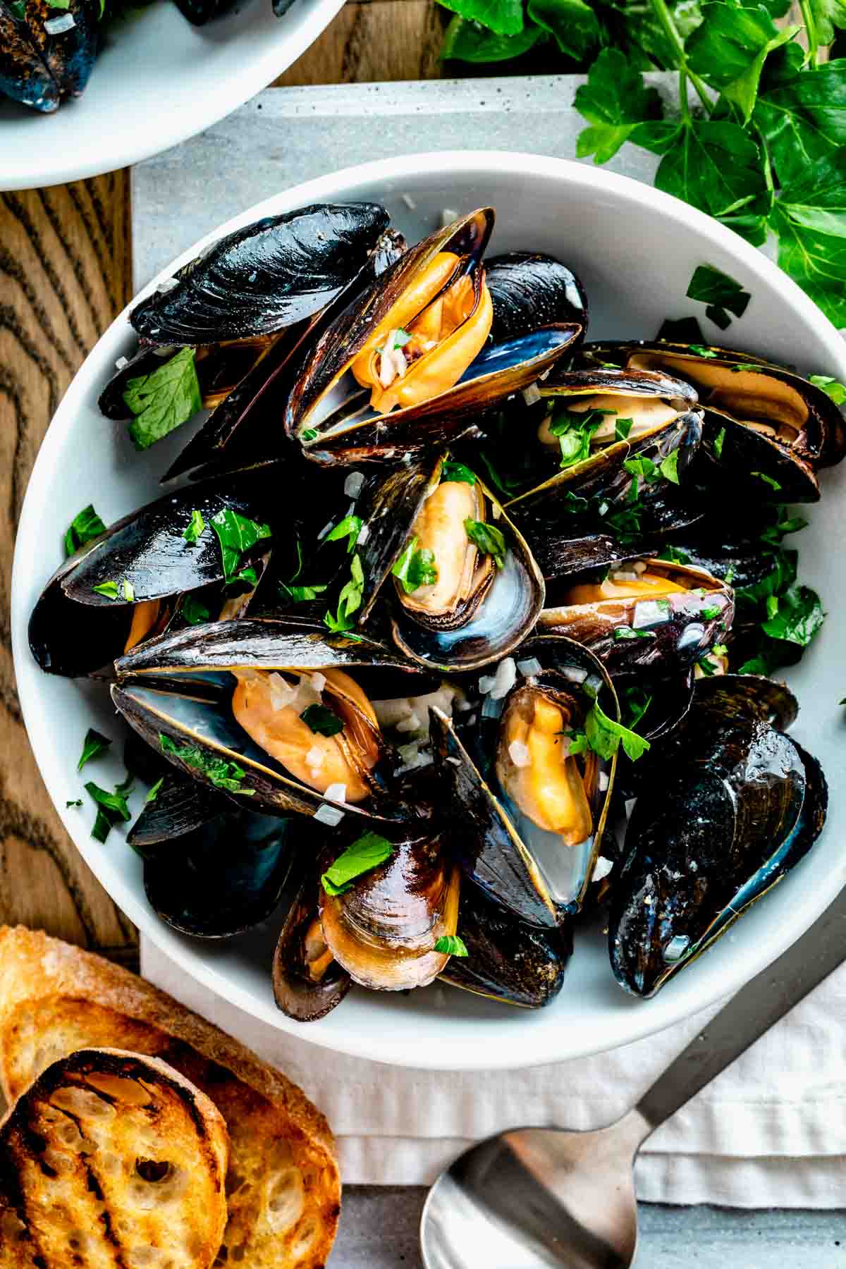 a white bowl filled with steamed mussels with parsley on top and two slices of grilled baguette on the table next to it