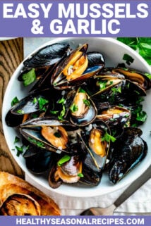 a white bowl filled with steamed mussels with parsley on top and two slices of grilled baguette on the table next to it