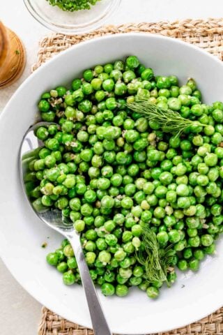 a white bowl with cooked peas, a spoon and sprigs of dill