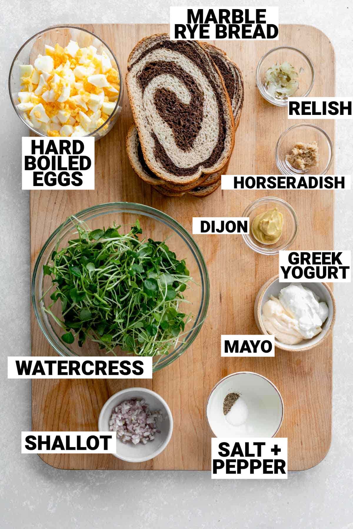 slices of marble rye, a bowl of watercress, chopped hard boiled eggs, and greek yogurt, shallot, salt and pepper, mayo, dijon, and horseradish laid out on a chopping board to make a watercress and egg salad sandwich
