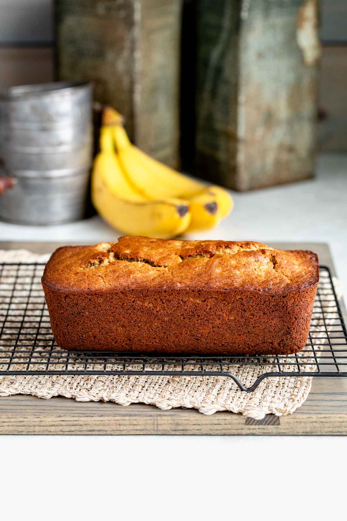 a loaf of white whole wheat flour banana bread on a wire rack cooling with bananas in the background