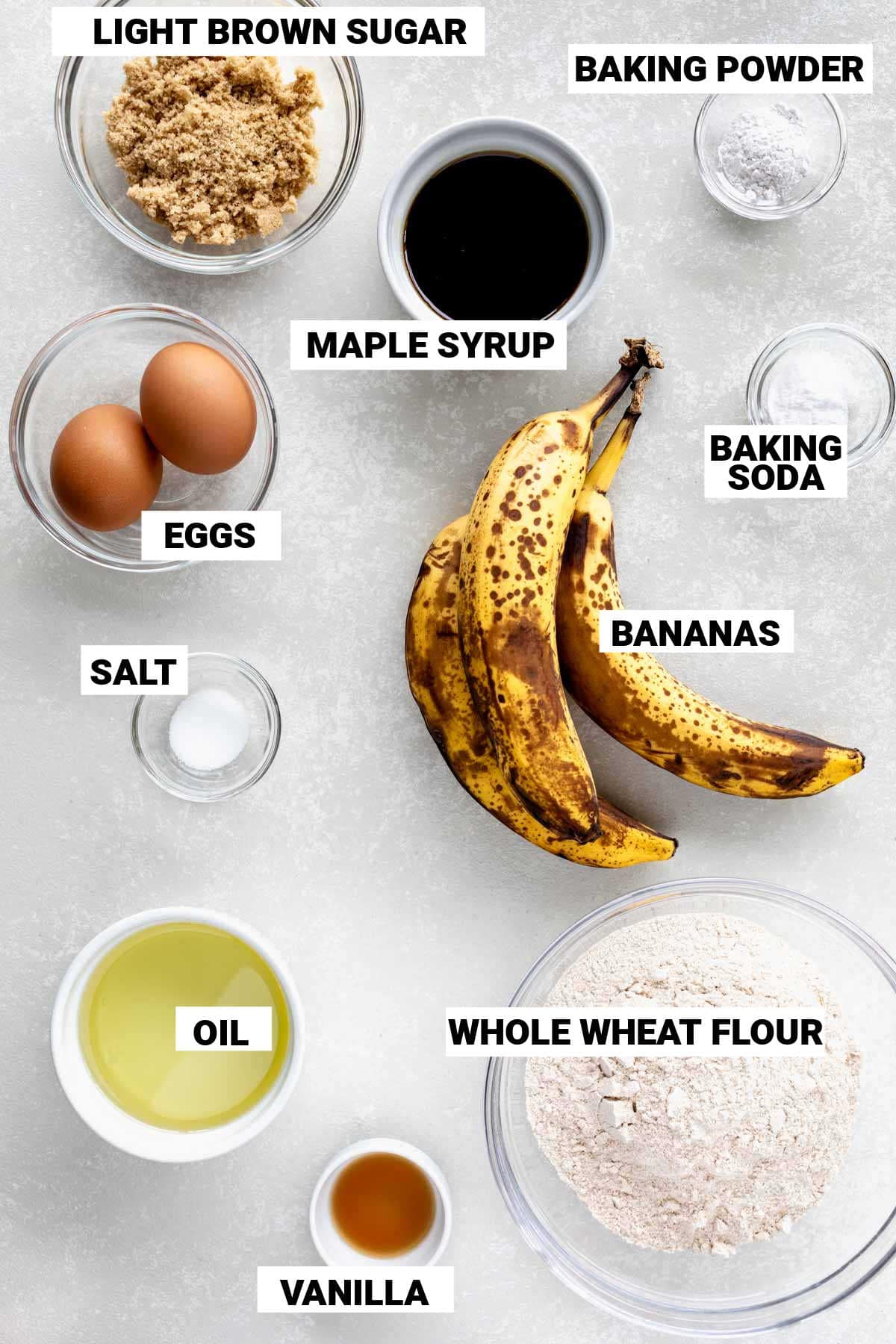 bananas, maple syrup, brown sugar, eggs, whole wheat flour, oil, baking soda and powder, vanilla, and salt laid out on a table to make whole wheat banana bread