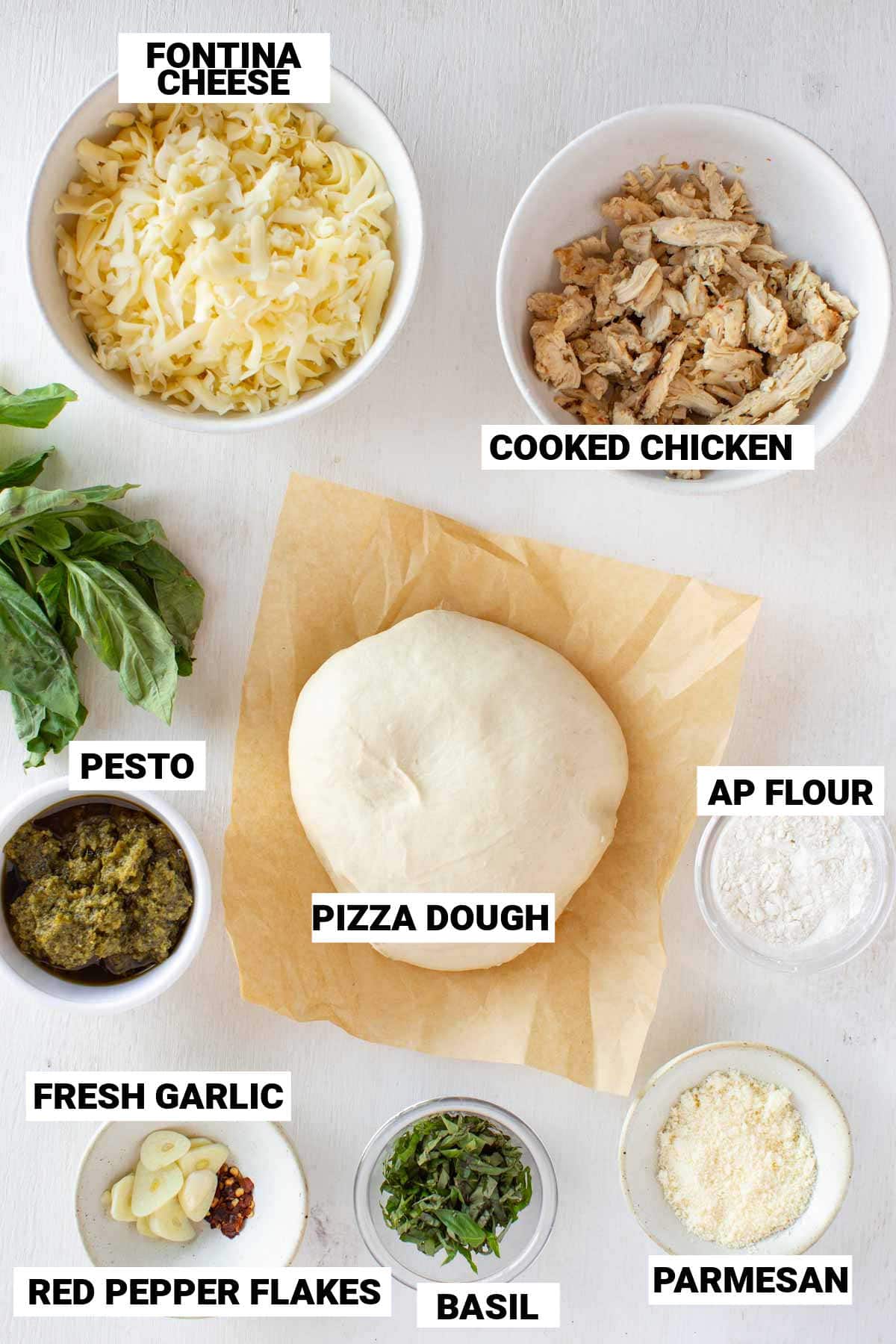 fontina cheese, cooked chicken, pizza dough, AP flour, pesto, fresh garlic, basil, parmesan, and red pepper flakes all laid out ready to make chicken pesto pizza