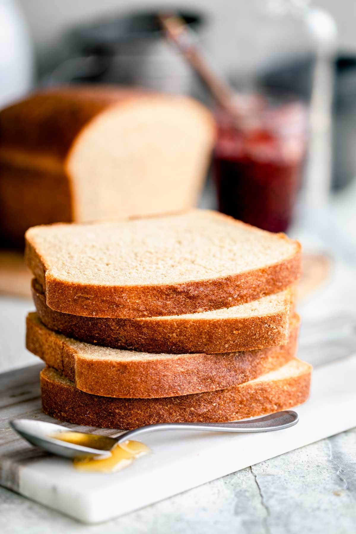 a stack of slices of honey whole wheat bread with a spoonful of honey in front of it and the rest of the loaf in the background