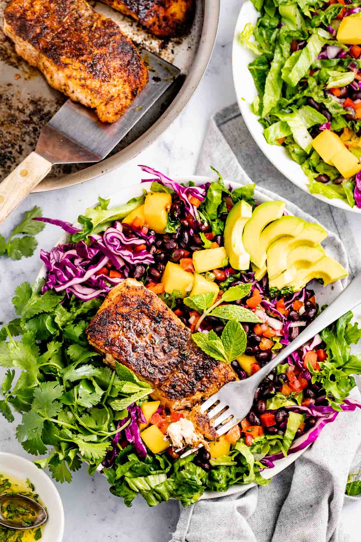 a large bowl of blackened salmon salad with the skillet with more salmon on it in the upper left corner and another bowl of salad to the right