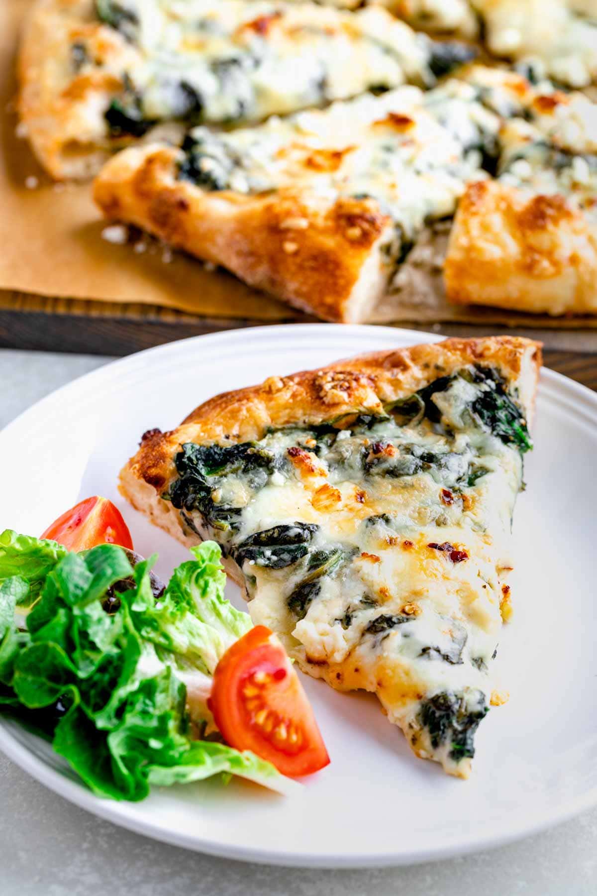 slice of homemade spinach pizza on circular white plate with a simple salad next to it.