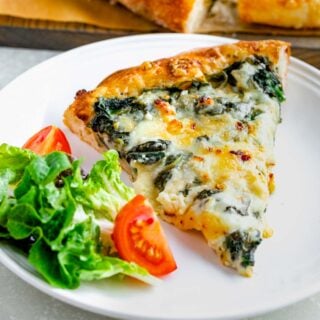 close up of triangle slice of homemade spinach pizza on circular white plate with simple salad next to slice