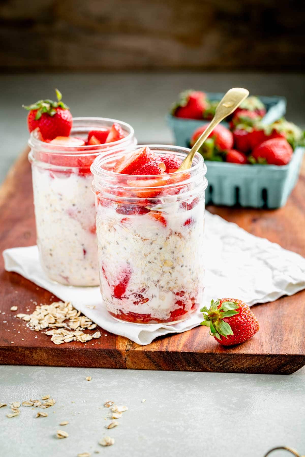 two mason jars full of strawberry overnight oats, with two small blue crates of strawberries in the background, and a strawberry and some oats scattered on the wooden table in the foreground