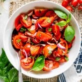 Tomato salad with balsamic vinaigrette in a white bowl from overhead with cherry tomatoes on the table and basil sprigs.