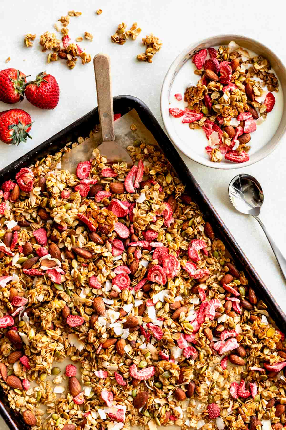 a sheet pan full of berry granola with a bowl of milk and granola to the side. Fresh strawberries and granola clusters are scattered around, along with a spoon.