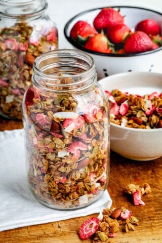 homemade strawberry granola in a couple of mason jars, with a bowl of milk and granola to the side and a bowl of fresh strawberries in the backround