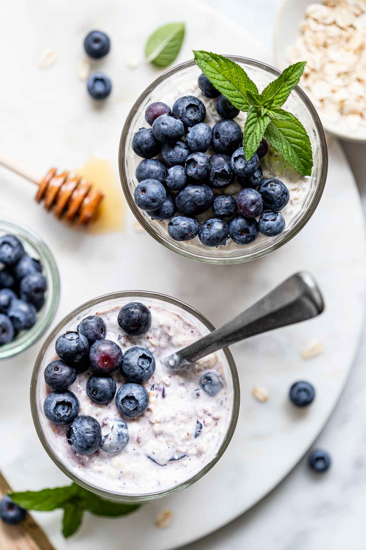 An overhead shot of fresh blueberries on top of overnight oats with green garnish and a spoon in one of the jars