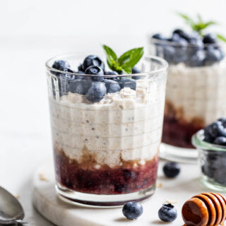 two cups of blueberry overnight oats with the mashed blueberries on the bottom, the oats layered on top, and fresh blueberries and garnish on the very top