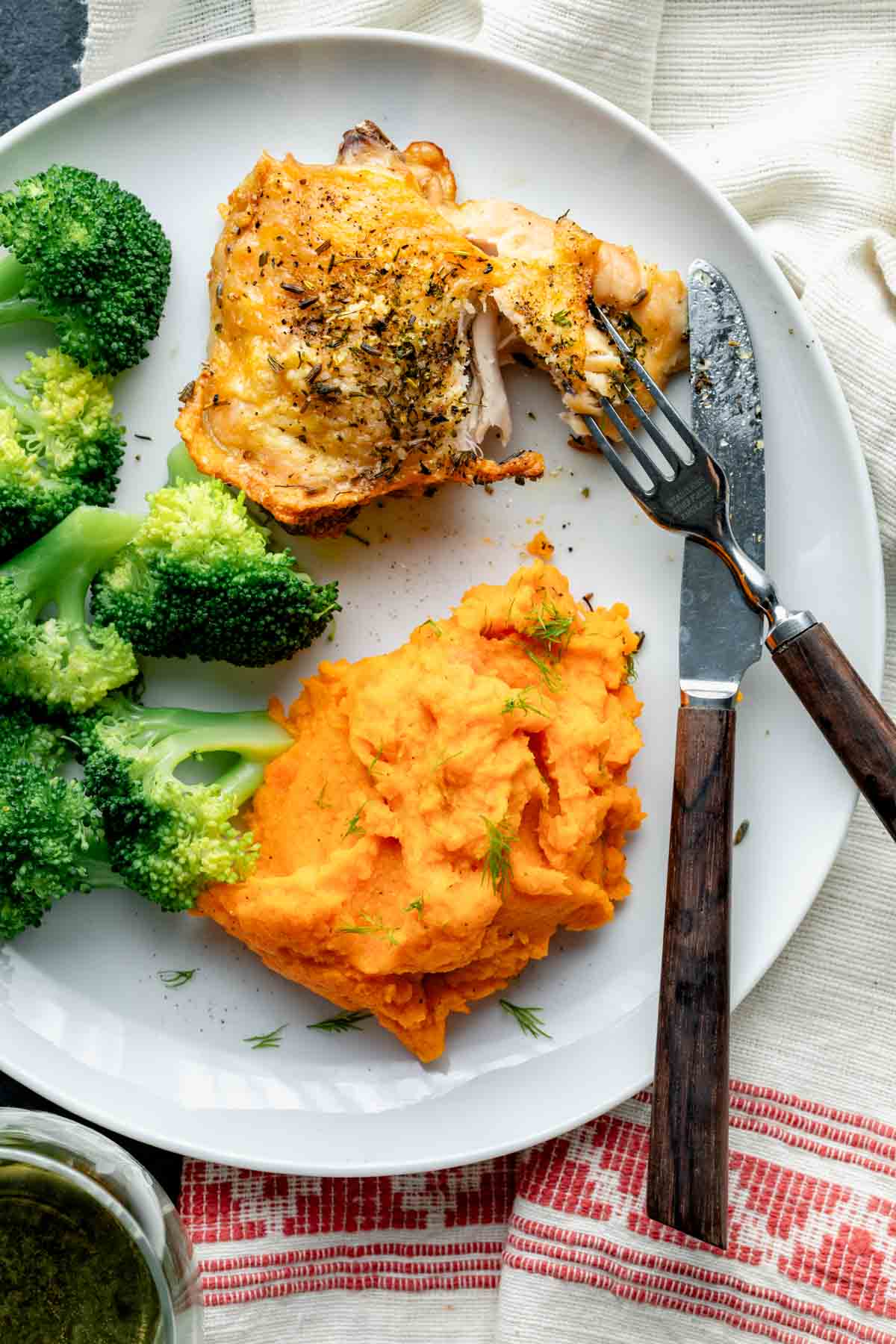 a baked chicken thigh on a plate with side dishes and a fork and knife cutting into it