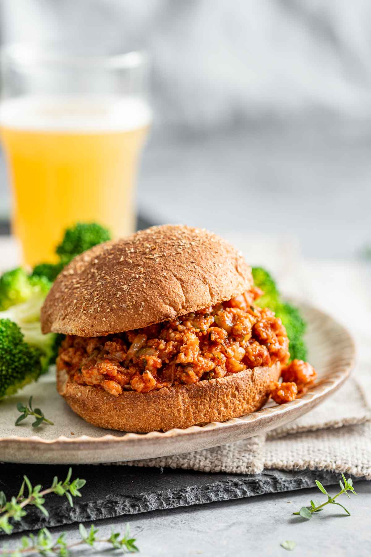 a healthy ground turkey sloppy jane on a bun sitting on a plate with broccoli and an orange beverage in the background