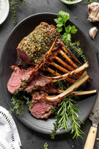 garlic and herb crusted lamb rib roast on a black oval platter with fresh herbs