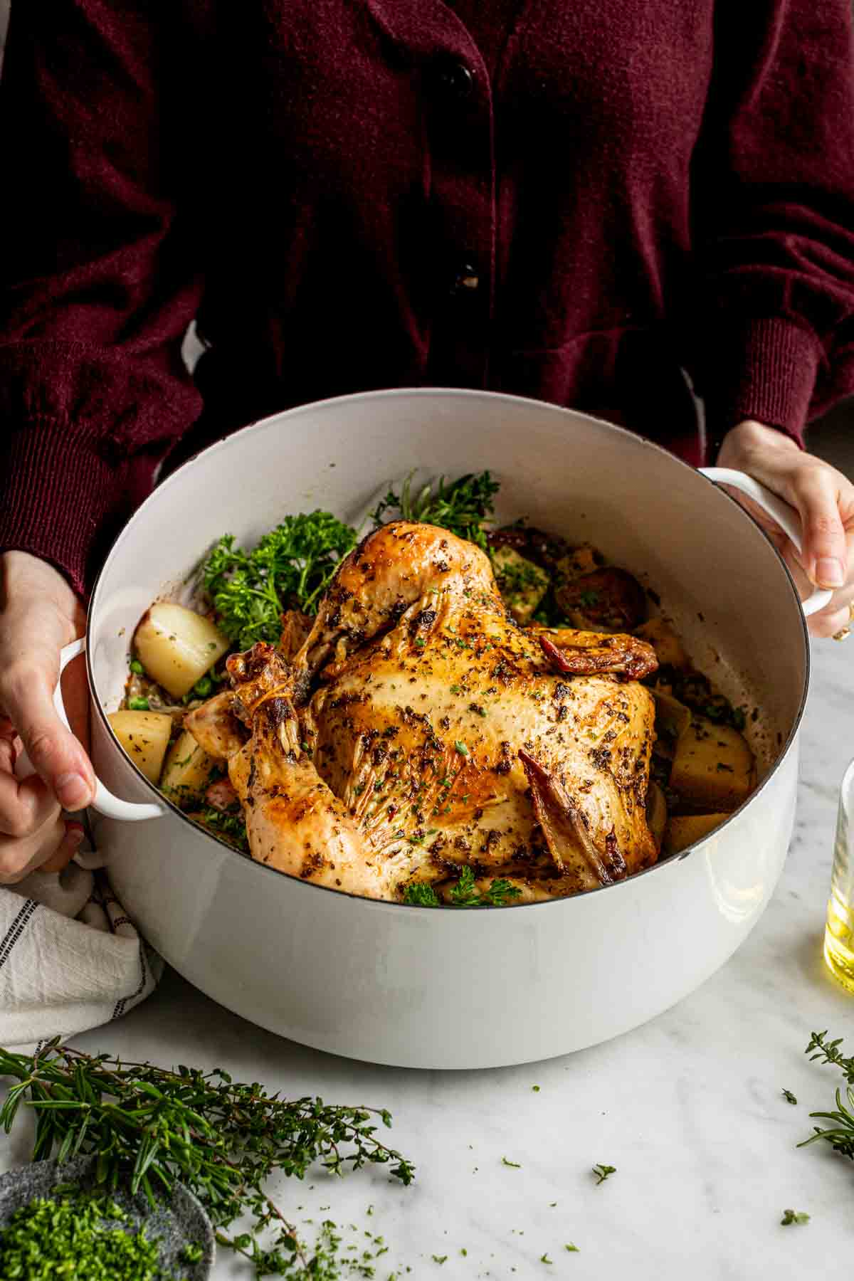 a person in a burgundy shirt holding the dutch oven to show the whole roast chicken and vegetables inside