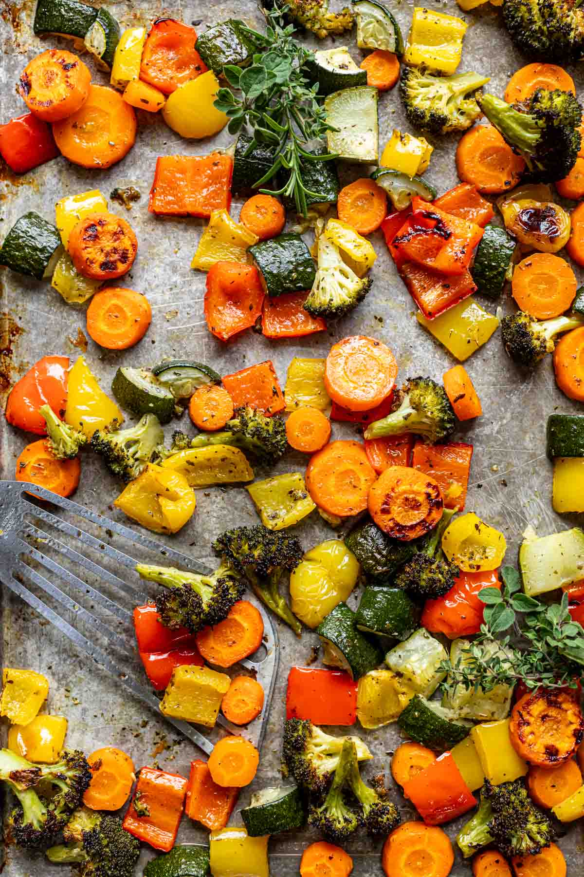 close up view of a variety of oven roasted veggies on a silver sheet pan with a spatula scooping some of the broccoli, bell peppers, and carrots