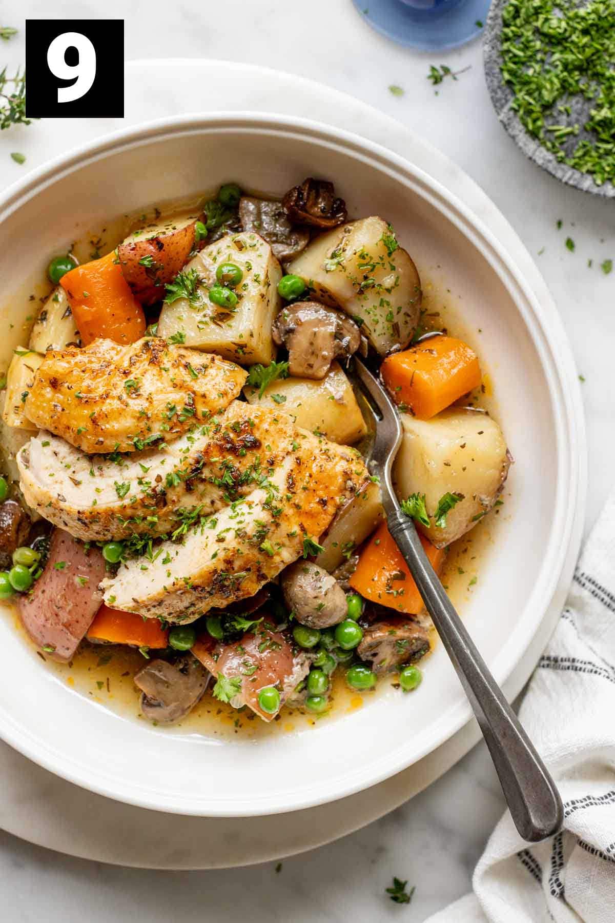 the roast chicken and vegetables in a white bowl with savory sauce and herbs for garnish, along with a fork sticking out of the dish