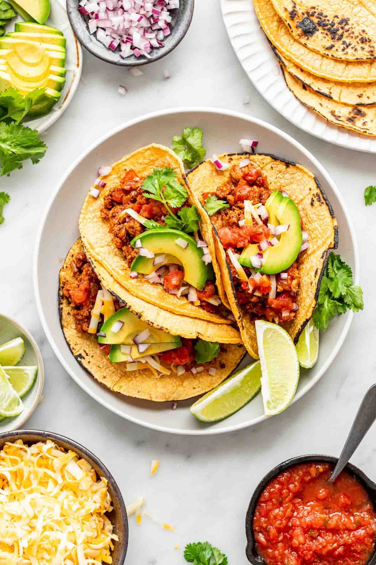 a white plate with three turkey tacos on it, topped with avocado slices, chopped red onion, parsley, and some shredded cheese.