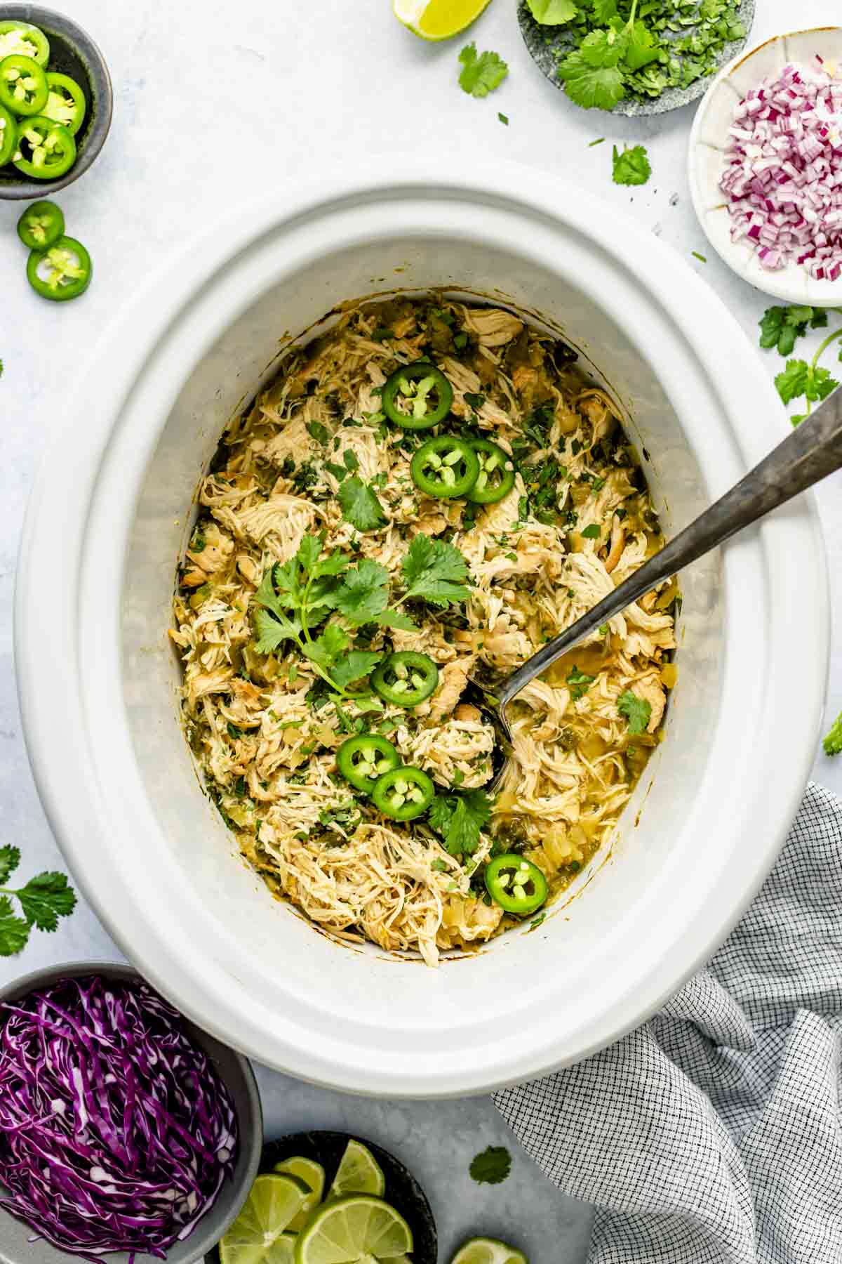 White Crockpot insert with shredded chicken, jalapenos and cilantro with a large silver serving spoon in it