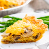 a slice of the mushroom quiche on a white plate with steamed asparagus and a shiny fork
