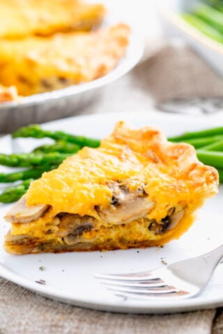 a slice of the mushroom quiche on a white plate with steamed asparagus and a shiny fork