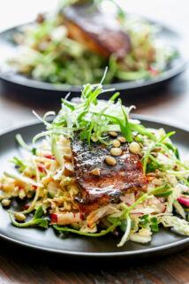 the blackened salmon on top of the shredded slaw with scallion frizzle on top on a black plate on a wooden tabletop