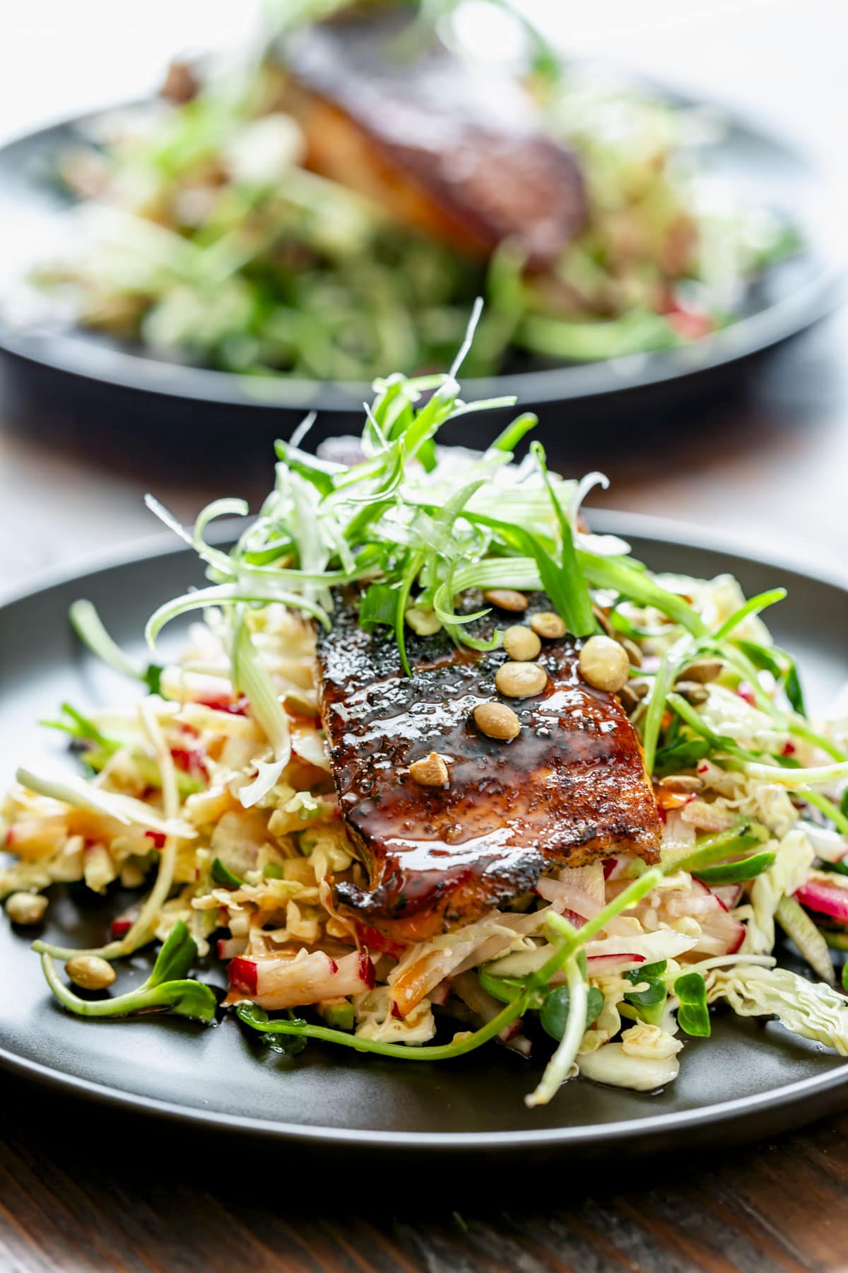 the blackened salmon on top of the shredded slaw with scallion frizzle on top on a black plate on a wooden tabletop