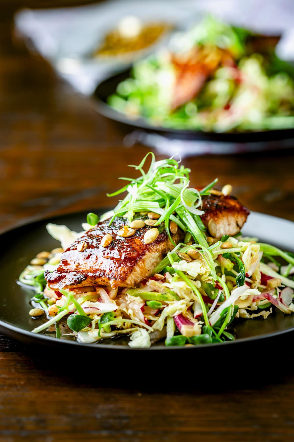 the blackened salmon on top of the shredded slaw with scallion frizzle on top on a black plate on a wooden tabletop