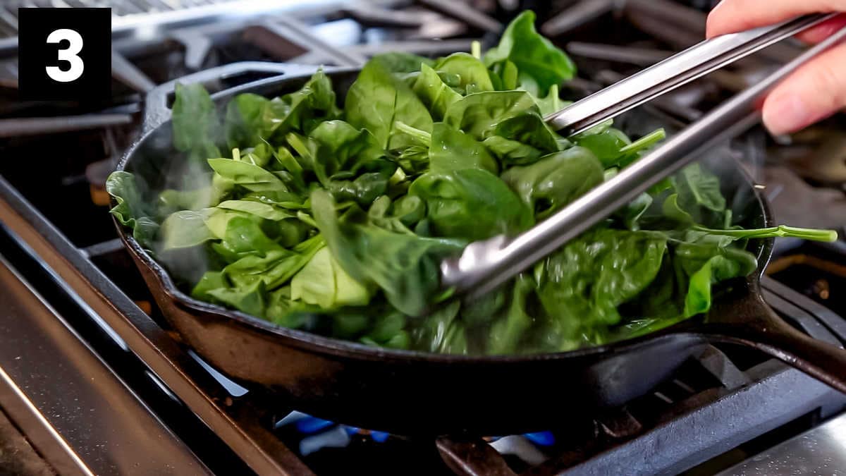 the cast iron skillet from the side, filled with spinach and a set of tongs reaching in to turn the spinach over