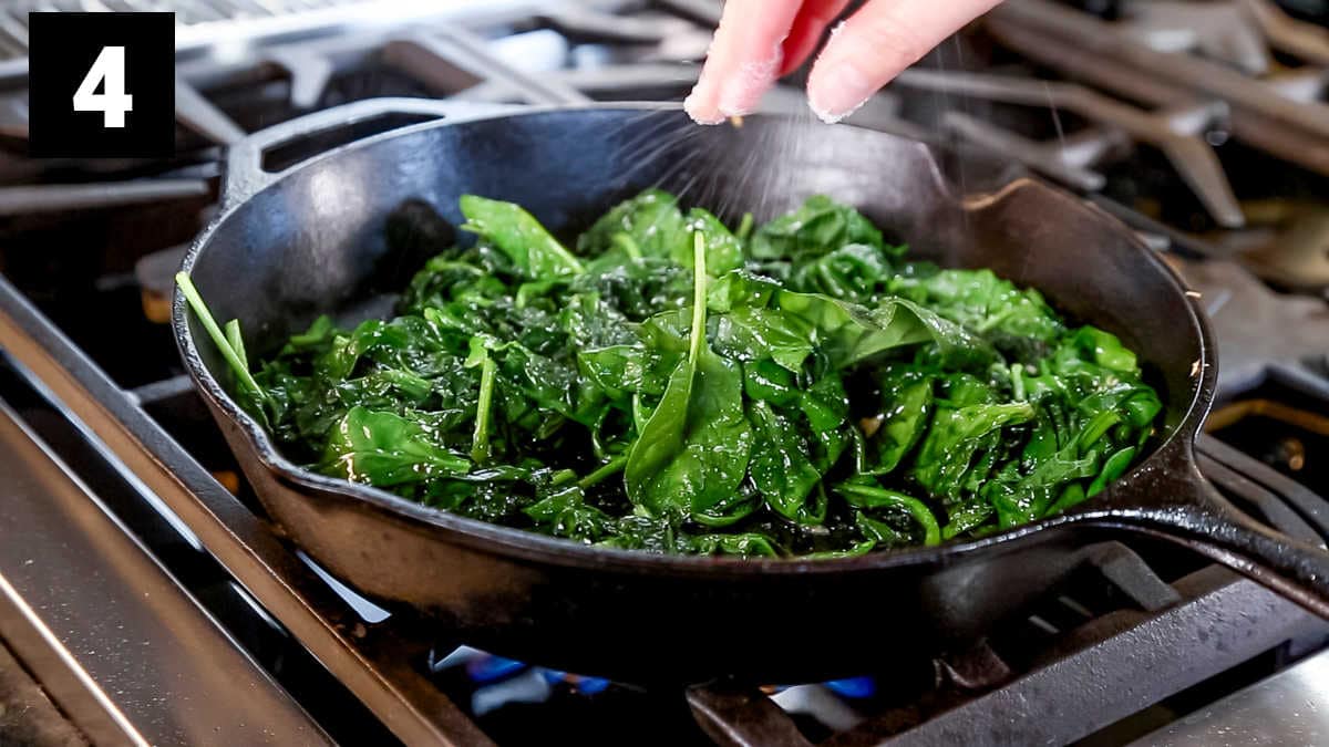 the cast iron skillet from the side with wilted spinach and fingers sprinkling coarse salt over it