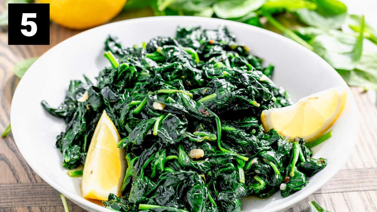 a white bowl filled with cooked baby spinach and lemon wedges