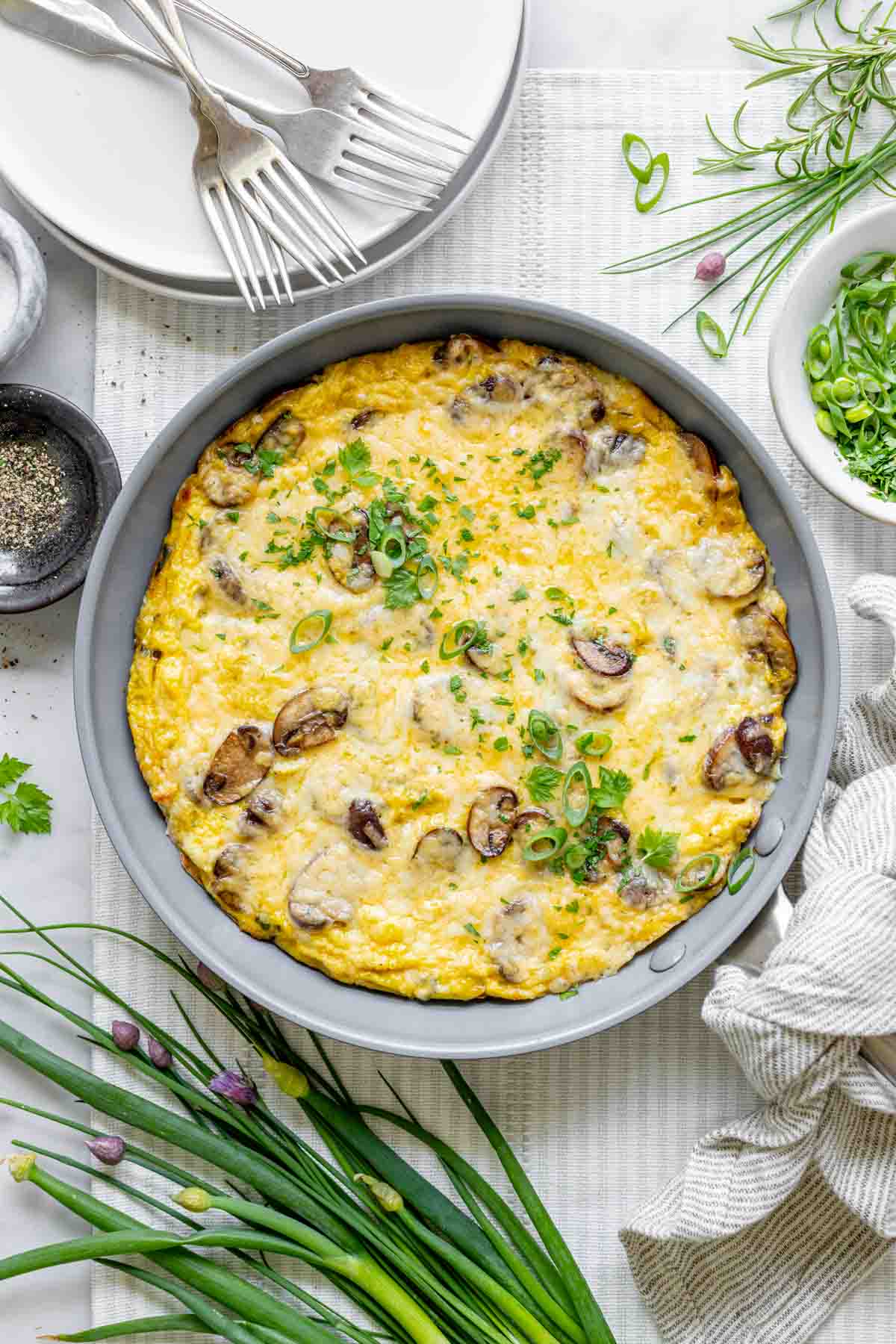 the frittata in the skillet from overhead with fresh herbs, a stack of plates and chive blossoms