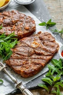 two pieces of lamb steak on a grey surface with fresh greens scattered around them